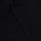 A close-up of the Abstract Jersey Cuba Shirt by 04651/A TRIP IN A BAG, a black lightweight cotton button-up with a pointed collar, three visible buttons, and a partially visible numbered white tag inside the neckline.