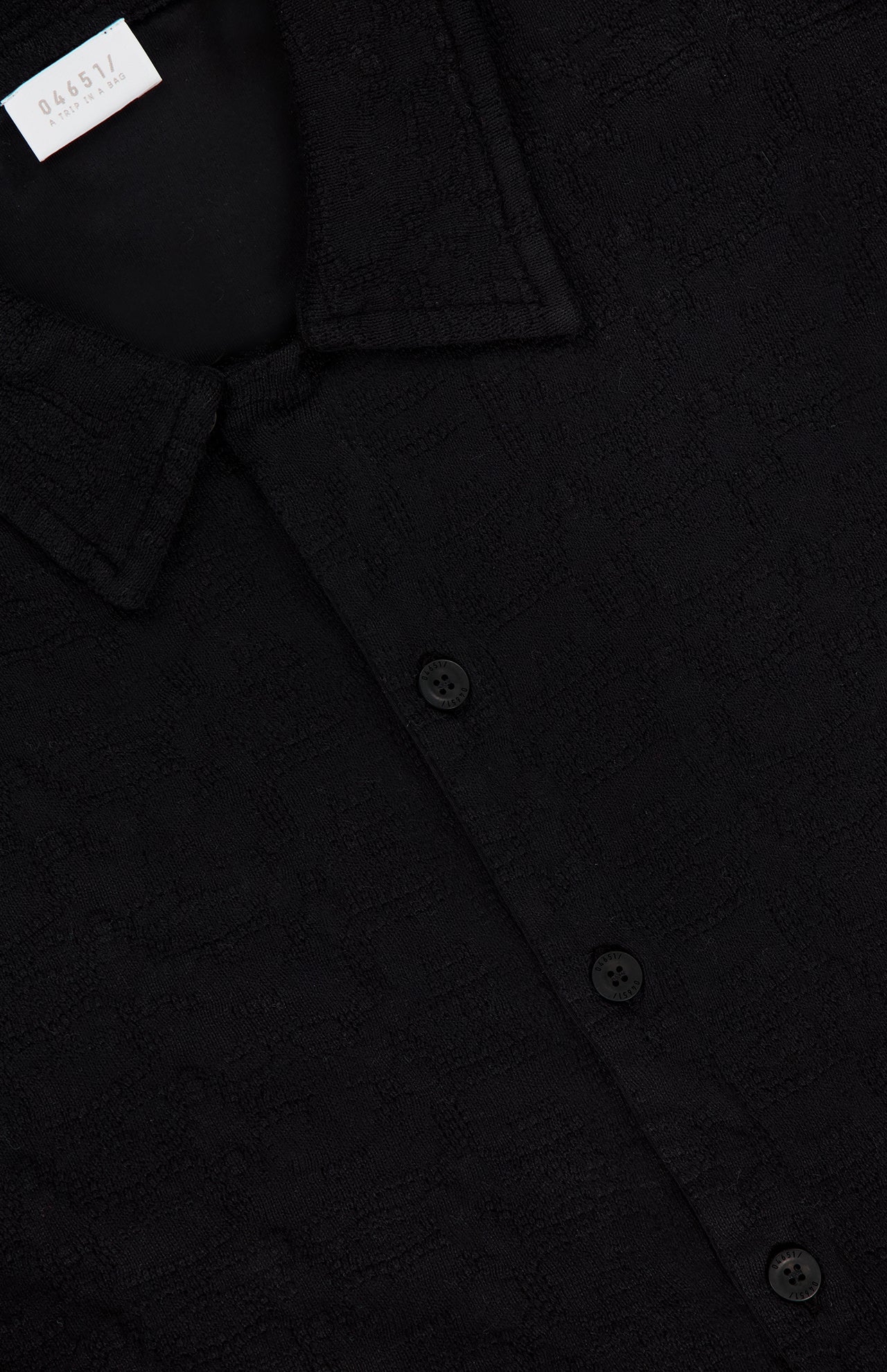 A close-up of the Abstract Jersey Cuba Shirt by 04651/A TRIP IN A BAG, a black lightweight cotton button-up with a pointed collar, three visible buttons, and a partially visible numbered white tag inside the neckline.