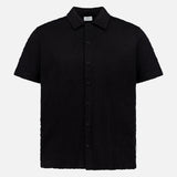 The Abstract Jersey Cuba Shirt by 04651/ A TRIP IN A BAG is a lightweight cotton, short-sleeve, button-up black shirt with a classic Cuban collar, shown against a plain white background.