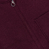 Close-up of the Boiled Virgin Wool Vest by 04651/ A TRIP IN A BAG in a maroon wine tone, featuring textured fabric, a pocket, and a circular zipper pull. A partially visible white tag with numbers appears at the top of the image.