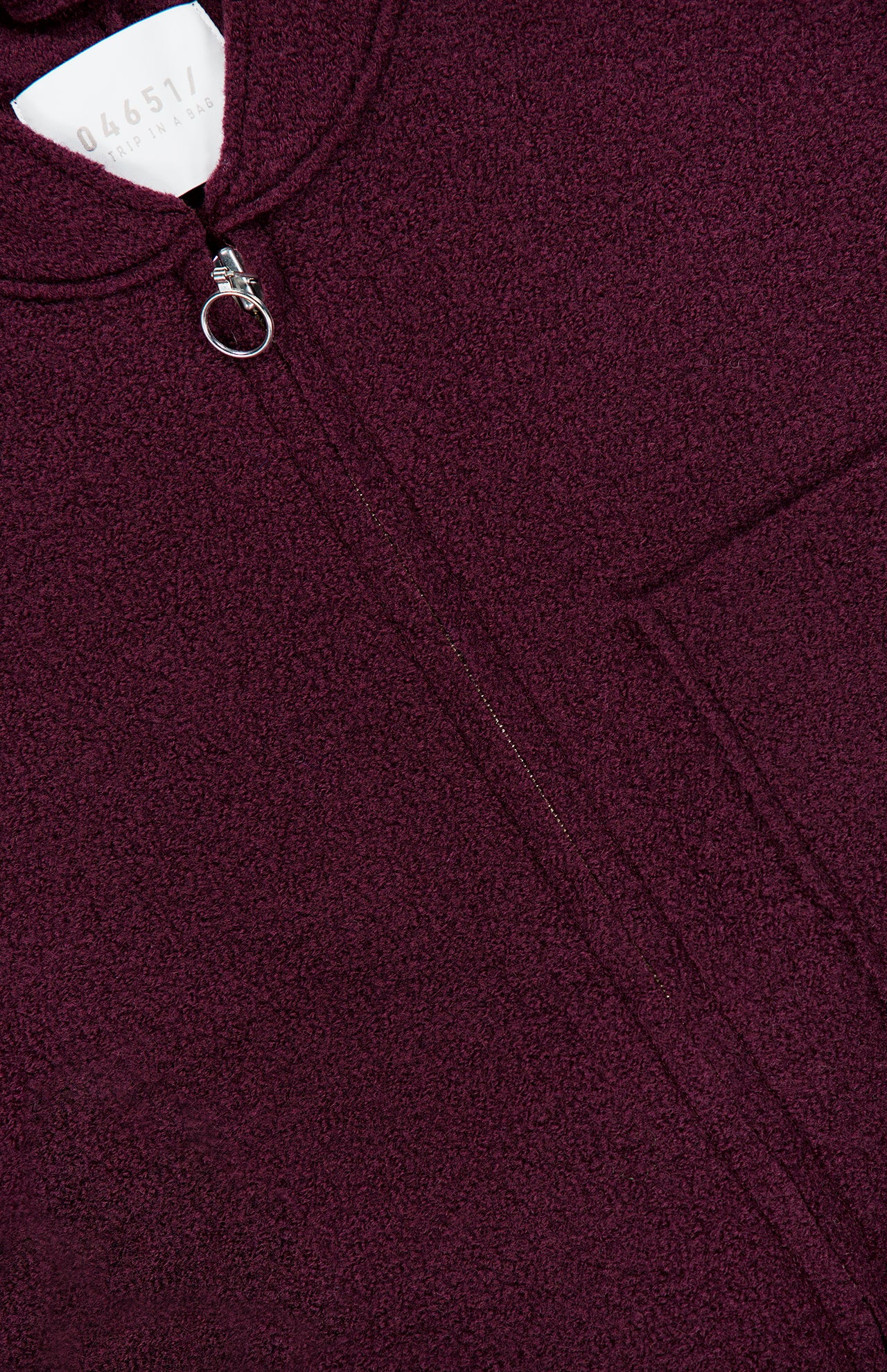 Close-up of the Boiled Virgin Wool Vest by 04651/ A TRIP IN A BAG in a maroon wine tone, featuring textured fabric, a pocket, and a circular zipper pull. A partially visible white tag with numbers appears at the top of the image.