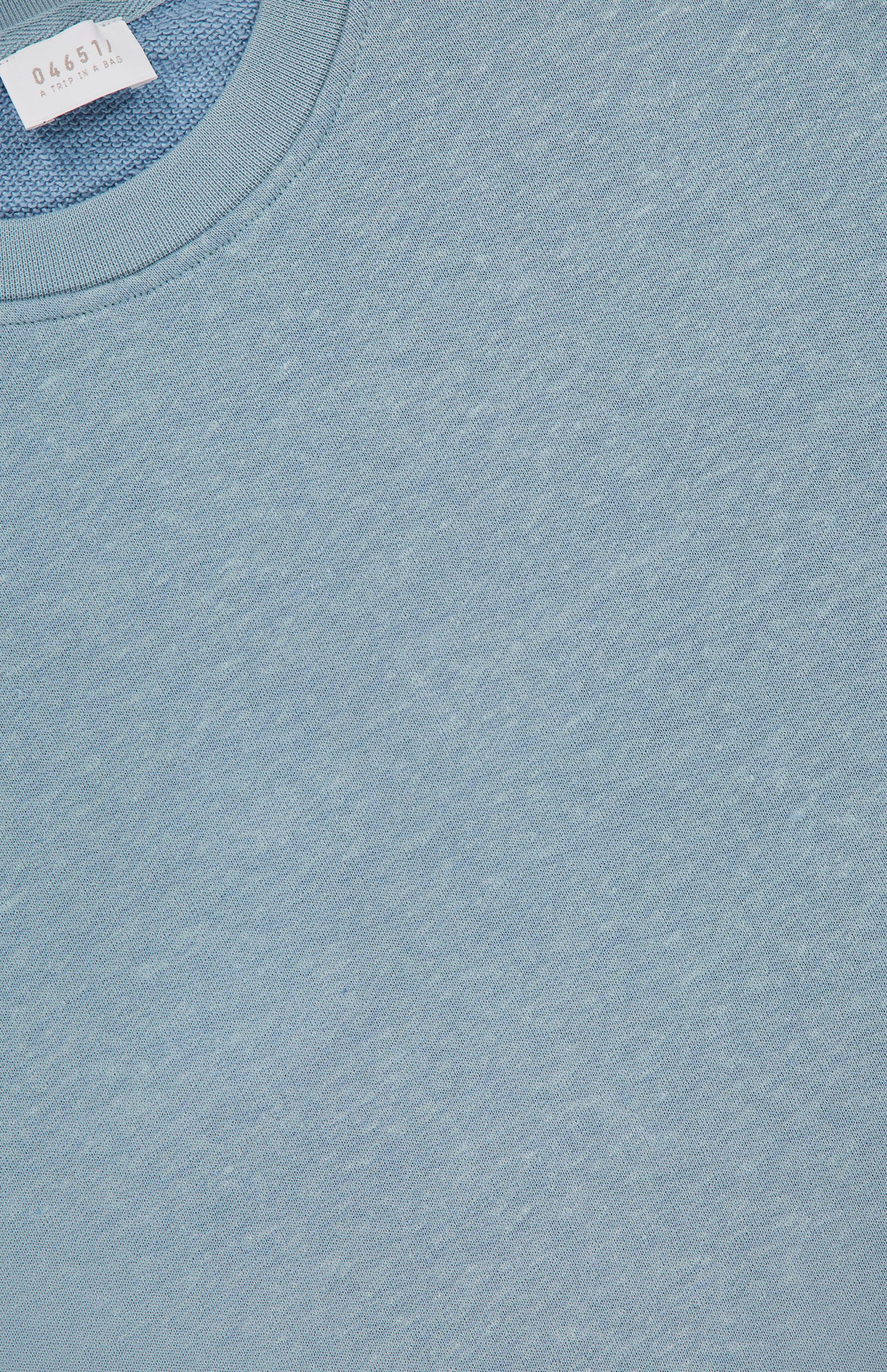 Close-up of the 04651/ A TRIP IN A BAG Cotton Linen Crewneck Sweatshirt in light blue, highlighting the fabric texture and a white collar tag with printed text.