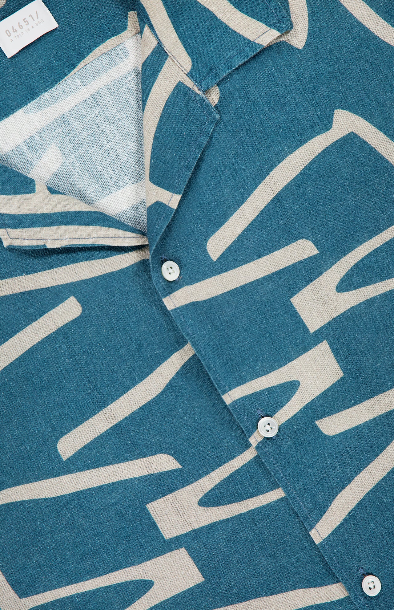 Close-up of the Hansen Retro Cuba Shirt by 04651/ A TRIP IN A BAG, featuring a teal and beige abstract patterned linen with a Cuban collar, white buttons, and a hint of retro design visible through the open side.