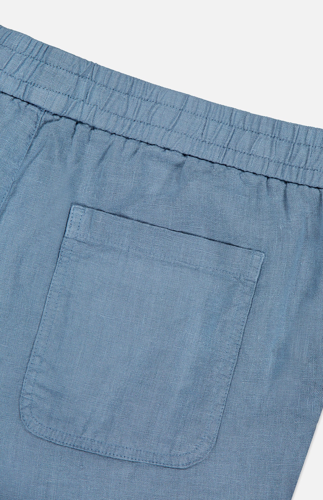 Close-up of the Linen Bermuda Short by 04651/ A TRIP IN A BAG, showing the back with a light blue linen fabric, elastic waistband, and single right patch pocket. Made in Portugal. Displayed against a plain white background.