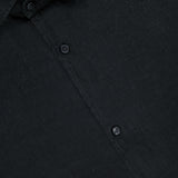 Close-up of the Linen Button-down Shirt by 04651/ A TRIP IN A BAG in black, showing its collar, three buttons, and a partial white tag; the men's casual shirt has slightly textured linen with a matte finish for a refined look.