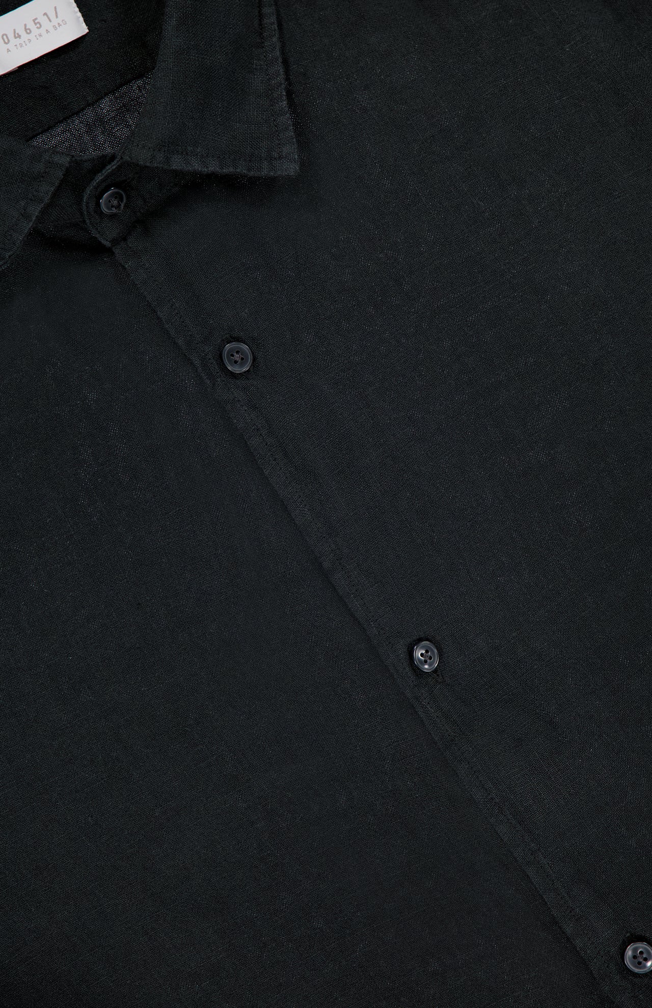 Close-up of the Linen Button-down Shirt by 04651/ A TRIP IN A BAG in black, showing its collar, three buttons, and a partial white tag; the men's casual shirt has slightly textured linen with a matte finish for a refined look.