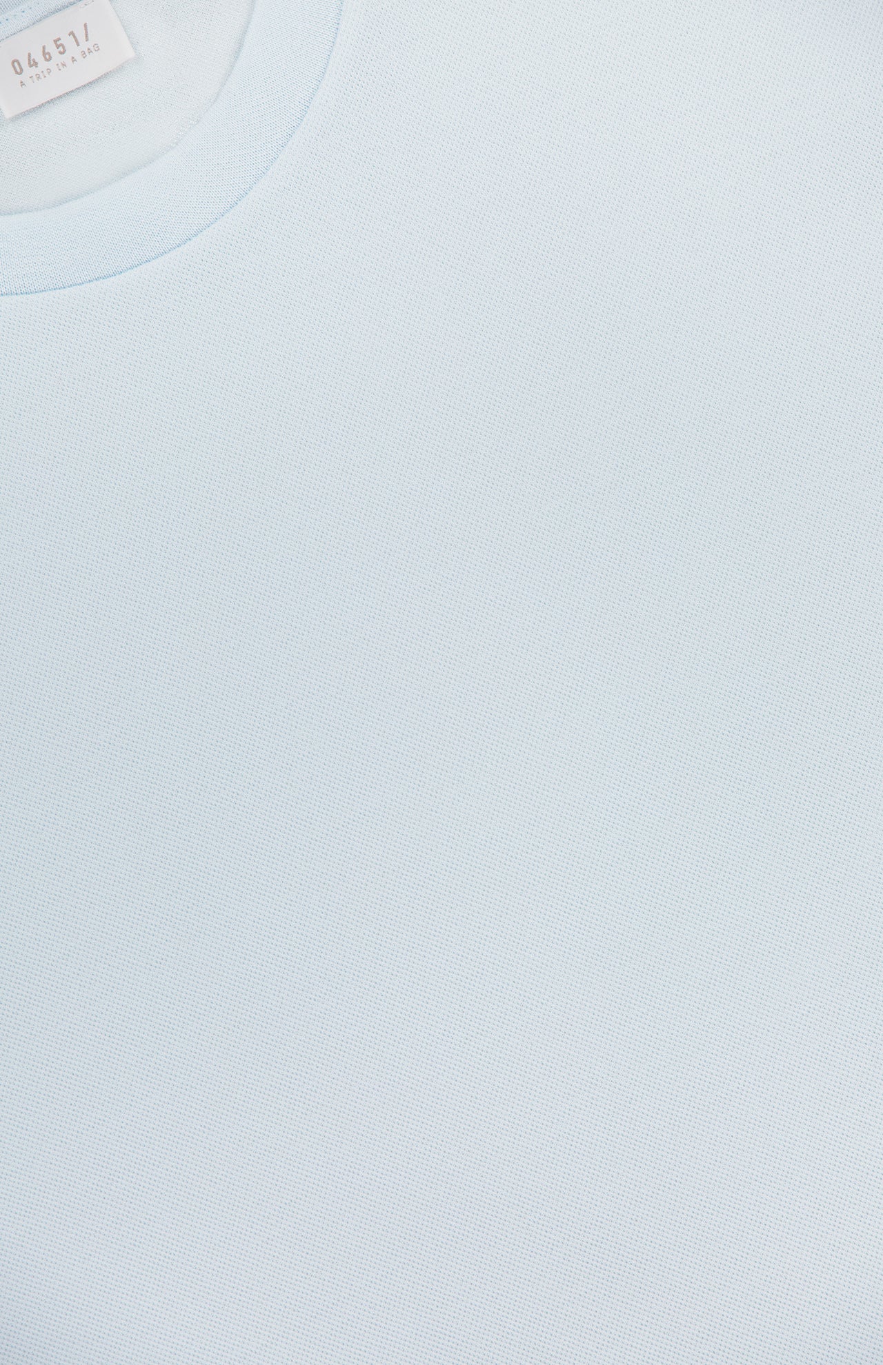 Close-up of the TP T-shirt by 04651/ A TRIP IN A BAG in light blue pure cotton, showing fabric texture and part of the neck label in the upper left corner.