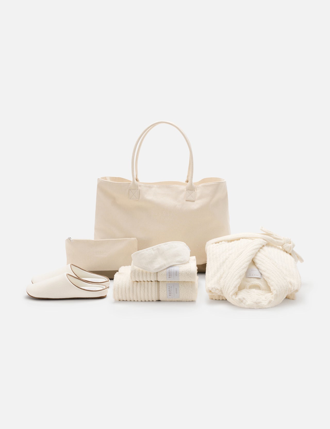 The Voyage Set Tote Bag by 04651/ A TRIP IN A BAG, along with a matching zip pouch, folded towels, cozy slippers, and a folded waffle robe tied with a bow, are arranged neatly on a white background.