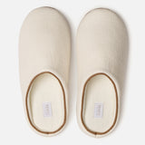 A pair of off-white, closed-toe slippers with a simple design, viewed from above. Both slippers have a soft texture and feature small white labels inside the heel area—making them a cozy travel essential to slip on after unpacking your Voyage Set Tote Bag by 04651/ A TRIP IN A BAG.