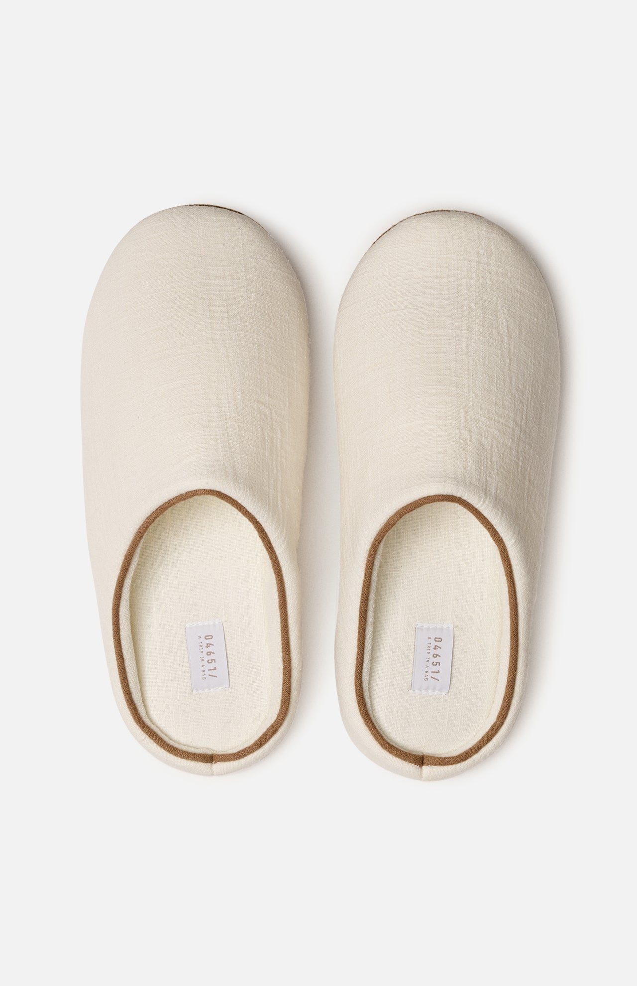 A pair of off-white, closed-toe slippers with a simple design, viewed from above. Both slippers have a soft texture and feature small white labels inside the heel area—making them a cozy travel essential to slip on after unpacking your Voyage Set Tote Bag by 04651/ A TRIP IN A BAG.