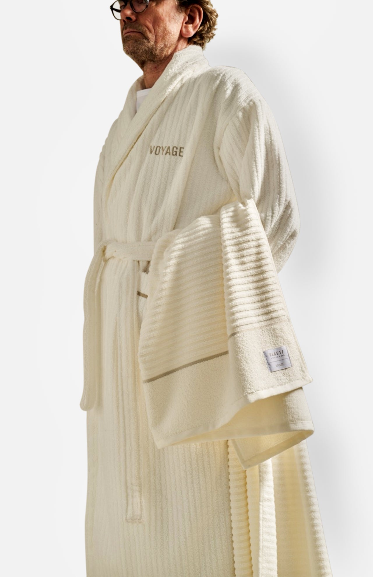 A man wearing glasses and a white textured bathrobe with “Voyage” embroidered on the chest holds the Voyage Set Tote Bag from 04651/ A TRIP IN A BAG—a true travel essential. The background is plain white.