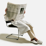 A person in a robe and slippers sits on a modern chair, holding and reading a large newspaper that covers their face and upper body, with the 04651/ A TRIP IN A BAG Voyage Set Tote Bag—an ideal travel tote—resting by their side against a plain light background.