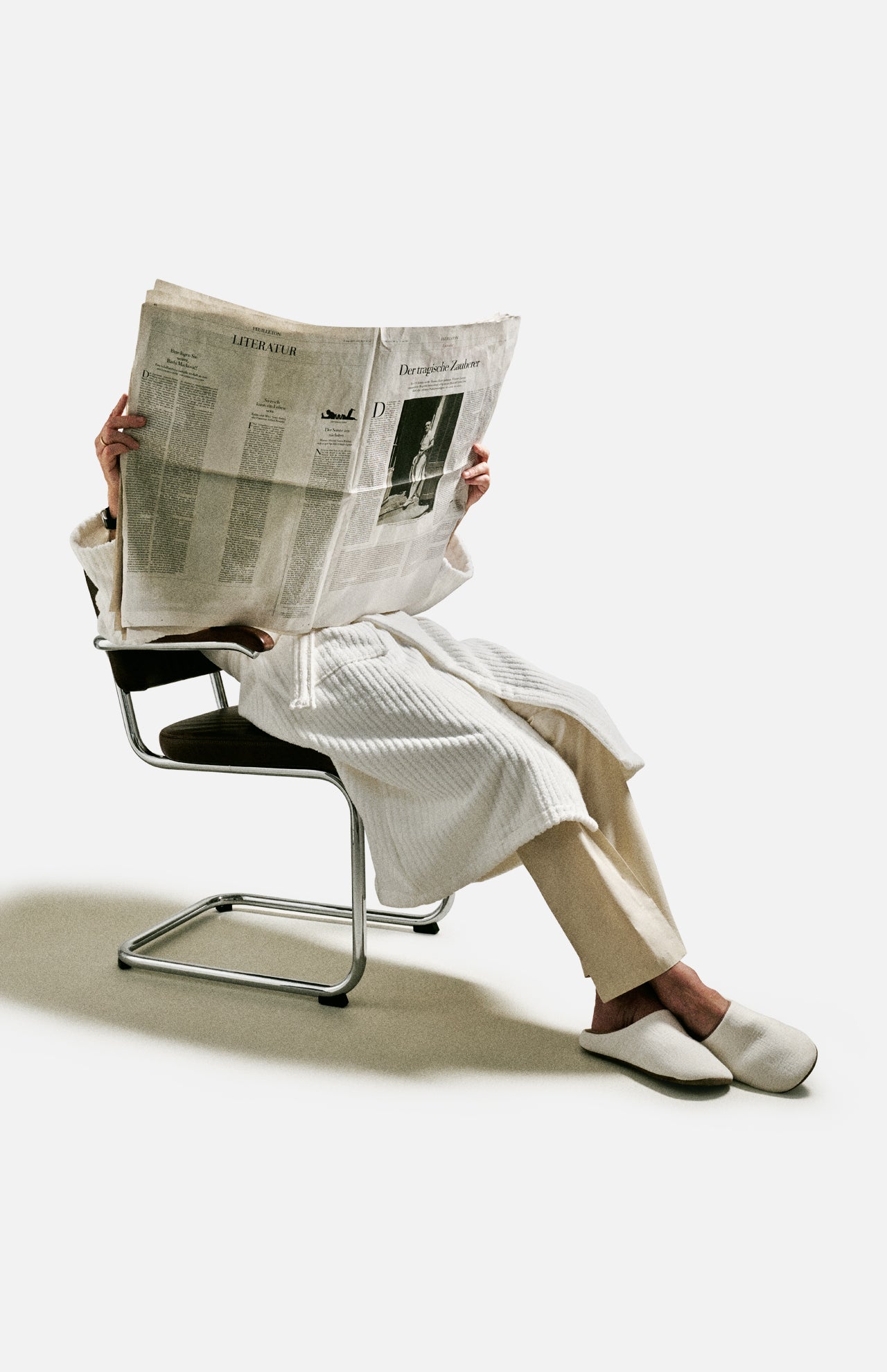A person in a robe and slippers sits on a modern chair, holding and reading a large newspaper that covers their face and upper body, with the 04651/ A TRIP IN A BAG Voyage Set Tote Bag—an ideal travel tote—resting by their side against a plain light background.