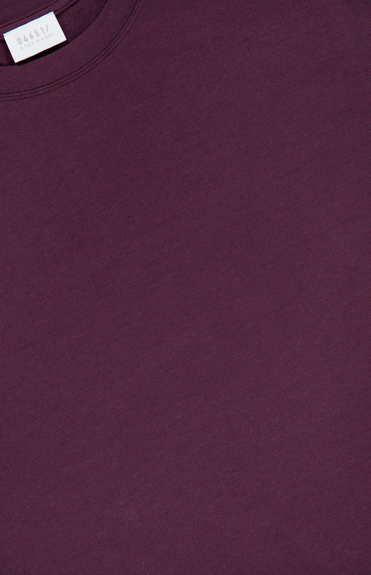 A close-up of the WJ Long Sleeve T-shirt by 04651/ A TRIP IN A BAG, shown in a plain, dark purple color with a small white clothing label visible at the neckline on the upper left side, made in Portugal.