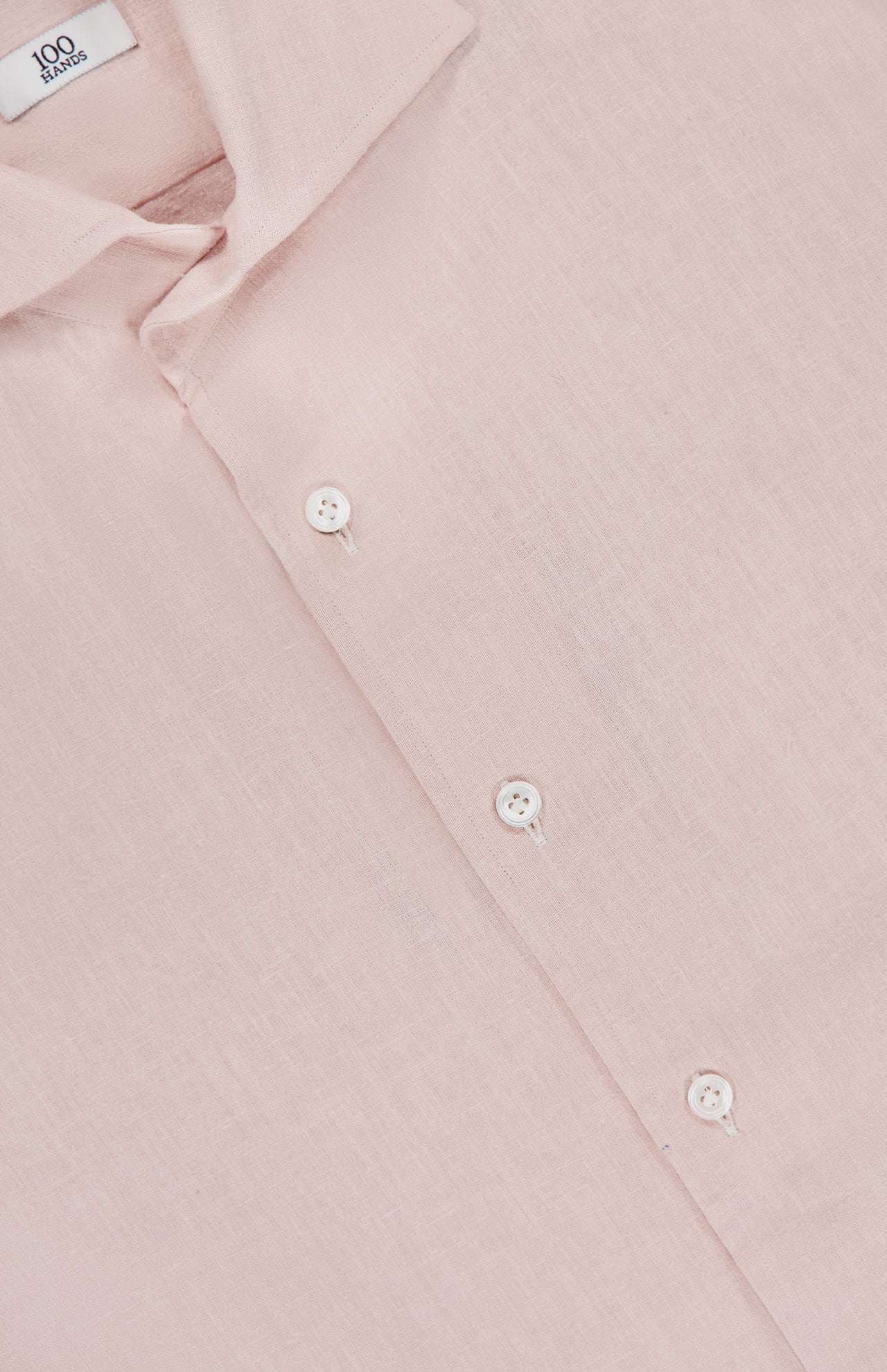 A close-up of the Canvas Dress Shirt by 100 HANDS, crafted in India from pale pink linen, features three white buttons and part of the collar, highlighting the shirt's soft, textured fabric.