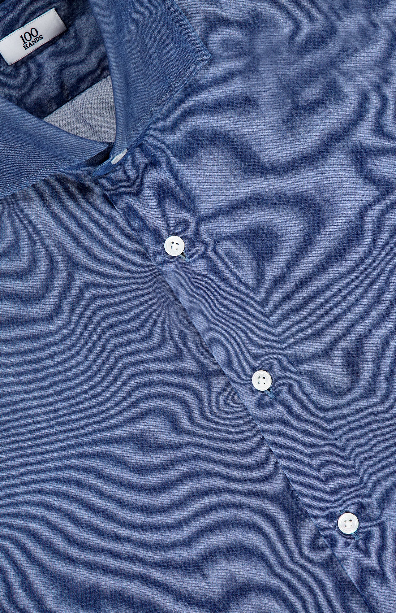 A close-up of the Denim Twill Shirt by 100 HANDS, a blue button-up made from lightweight fabric with white buttons and a collar. The label reads "100 HAND," emphasizing the artisanal quality of the brand.