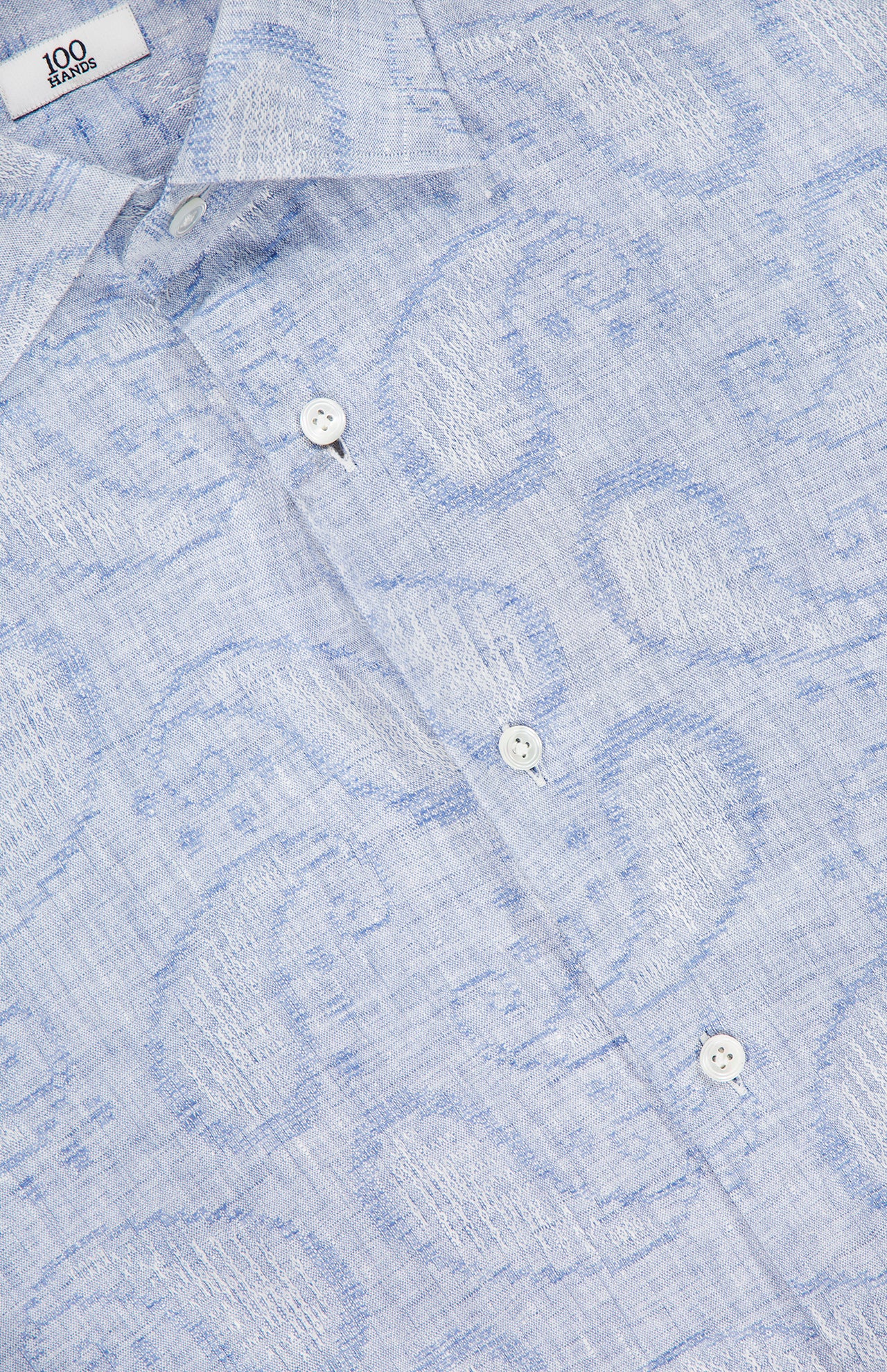 Close-up of the 100 HANDS Jacquard Paisley Dress Shirt in light blue, featuring a subtle paisley pattern, white buttons, and a visible collar with the signature label. The textured linen adds to its refined appeal.
