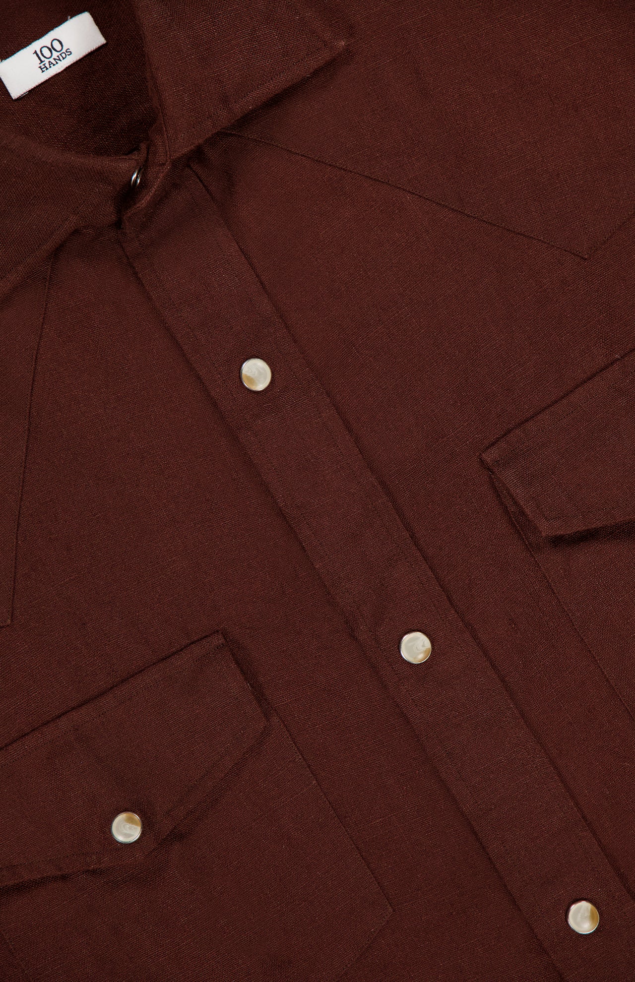 Close-up of a brown Linen Western Snap Shirt by 100 HANDS, featuring two front chest pockets, white snap buttons, and visible stitching. The collar and part of the 100 HANDS label are shown. Expertly crafted and made in India.