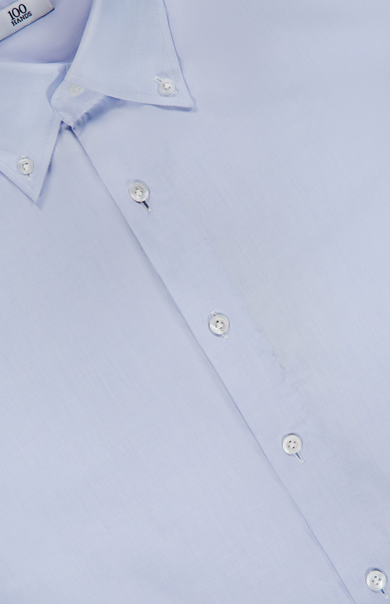 Close-up of the 100 HANDS Oxford Casual Fit Shirt in light blue cotton, featuring white buttons. The shirt is neatly pressed and laid flat, displaying its smooth yet subtly textured fabric. A visible collar and part of the shirt label are shown in the top left corner. This handmade piece exemplifies 100 HANDS’s commitment to craftsmanship and meticulous attention to detail.