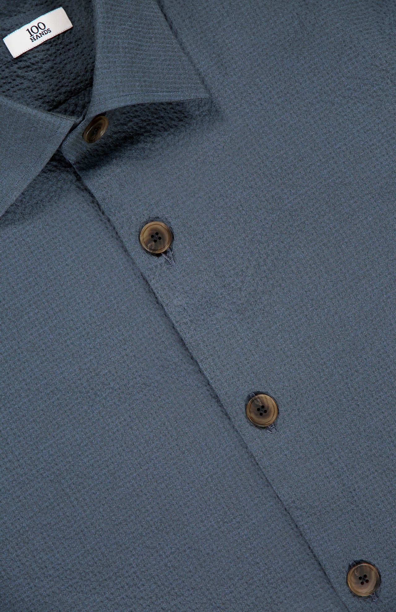Close-up of the 100 HANDS Seersucker Dress Shirt in textured blue fabric, brown buttons, and a white collar label with "100". The shirt is neatly pressed and has subtle patterns, showcasing quality craftsmanship from India.