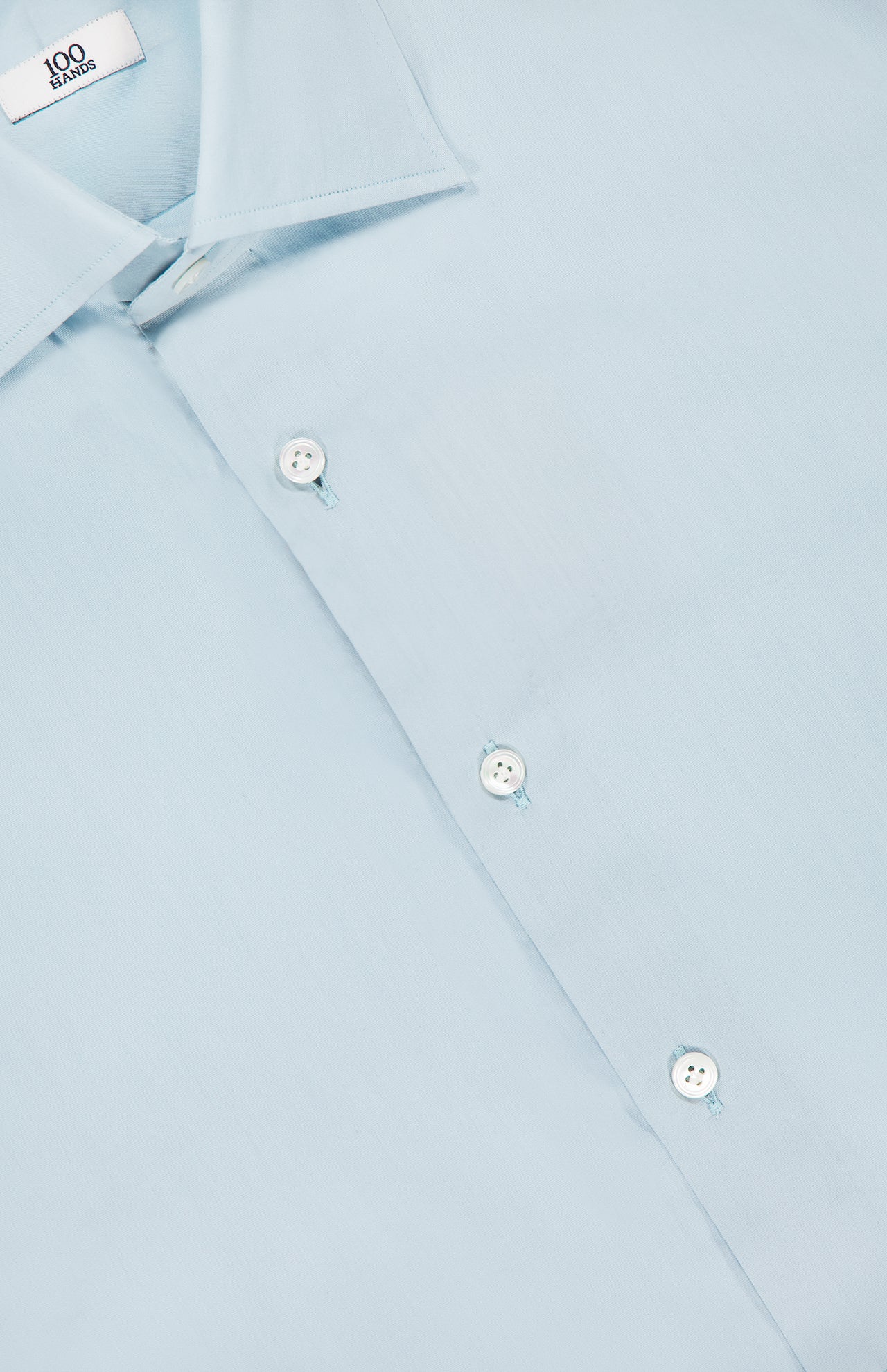 A close-up of a light blue Silk Twill Dress Shirt by 100 HANDS shows three white buttons, part of the collar, and a fabric label inside reading 100 HANDS, emphasizing its Indian craftsmanship.