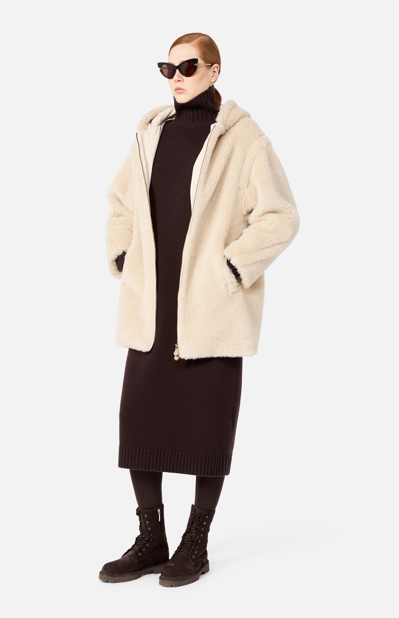 A person wearing a beige Max Mara Teddy Hooded Coat over a dark brown sweater dress, dark tights, and lace-up boots, with dark sunglasses.