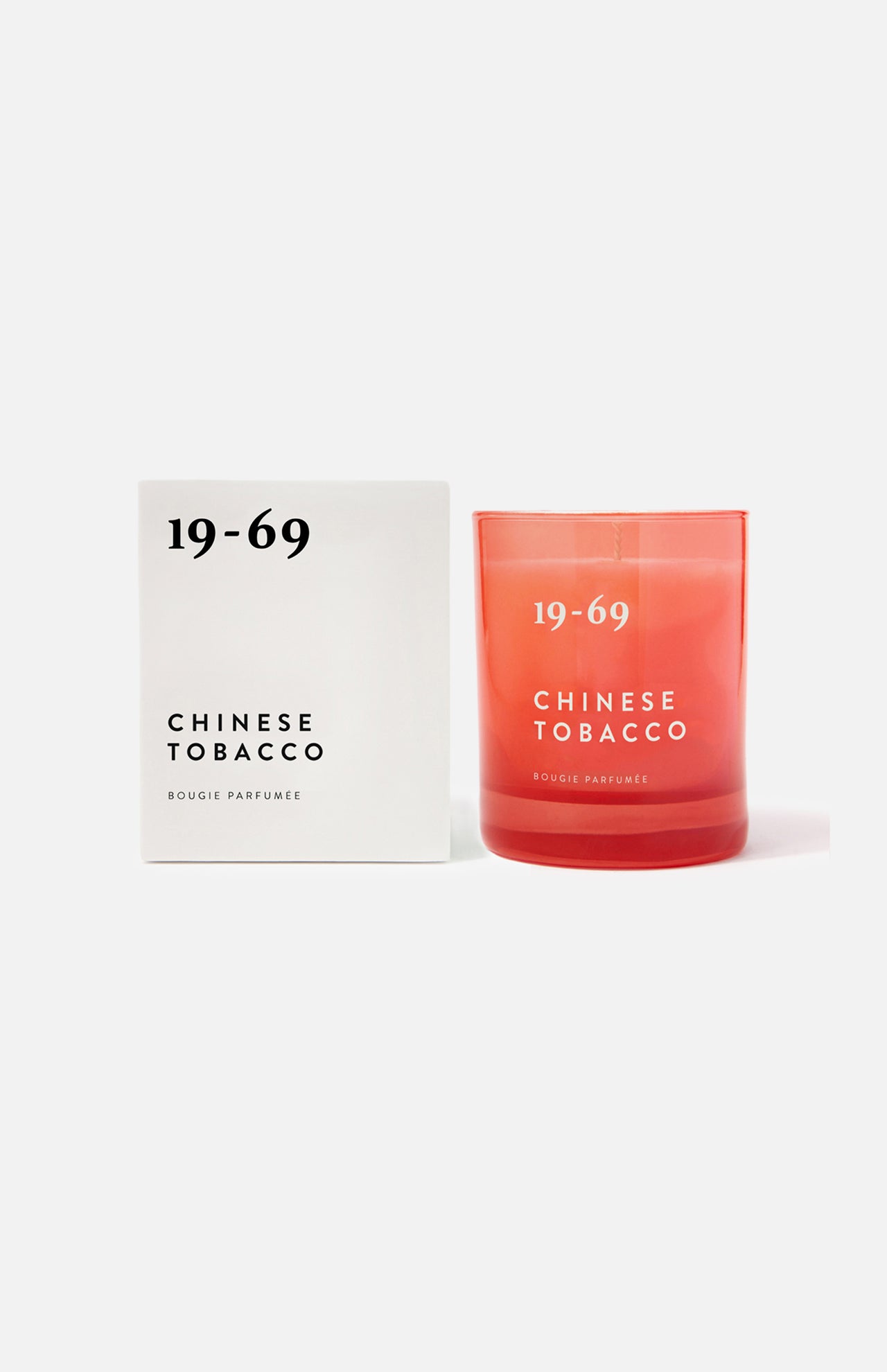 A white box labeled 19-69 Chinese Tobacco Candle stands beside a red glass candle holder, both set against a plain background. This unisex candle features a spicy woody scent inspired by Kuala Terengganu fragrance.