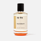 A clear glass bottle of 19-69 Kasbah EDP 100 ml Fragrance, a unisex scent with oriental spices and sandalwood, features light amber liquid, a black cap, and minimalist black text on a cream label against a plain white background.
