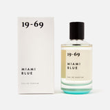 A bottle of 19-69 Miami Blu EDP 100 ml Fragrance stands beside its matching white box with black text on a plain white background. The unisex perfume has a black cap and contains clear liquid inside.