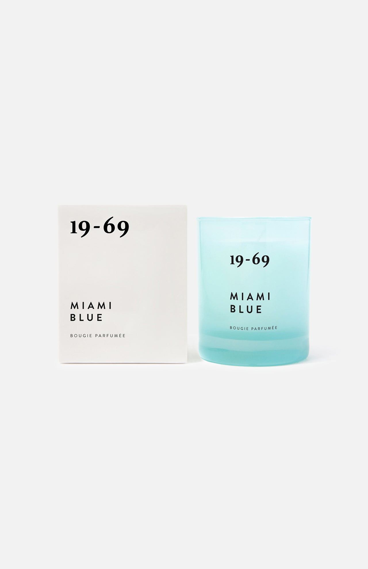 A light blue unisex Miami Blue Candle by 19-69 sits in a glass holder labeled with the brand and product name, next to a matching white box, against a plain background. It features a fresh aquatic scent inspired by Miami.