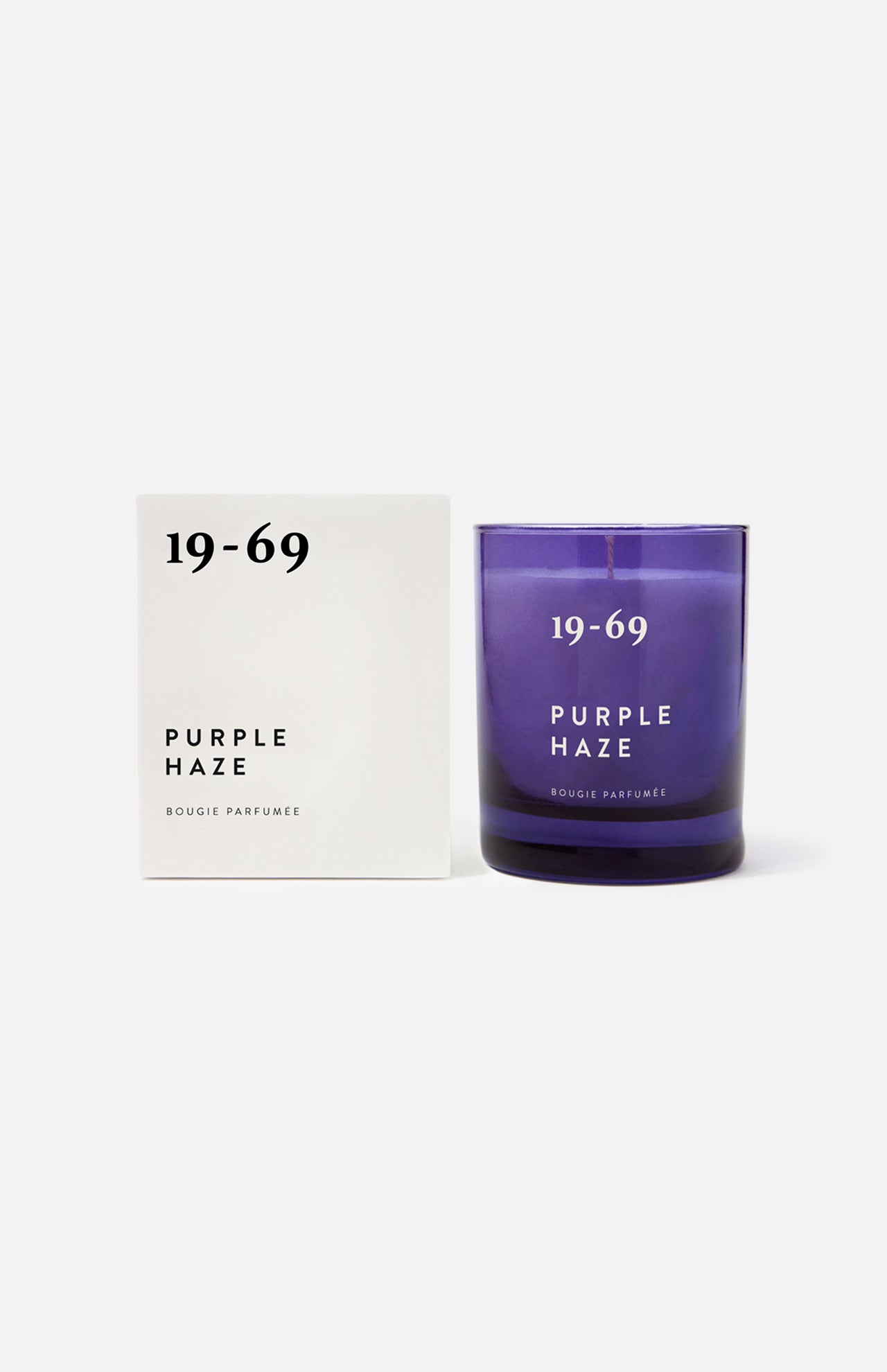 A white box labeled 19-69 Purple Haze Candle stands beside a matching purple glass candle, both set against a plain white background—a Woodstock-inspired scent featuring Italian bergamot from the brand 19-69.