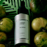 A silver bottle labeled “FLAMINGO ESTATE Roma Heirloom Tomato Room Spray” rests among green and yellow heirloom tomatoes in a green basket, evoking the essence of this room spray that captures the fresh, summer garden scent for your home fragrance collection.