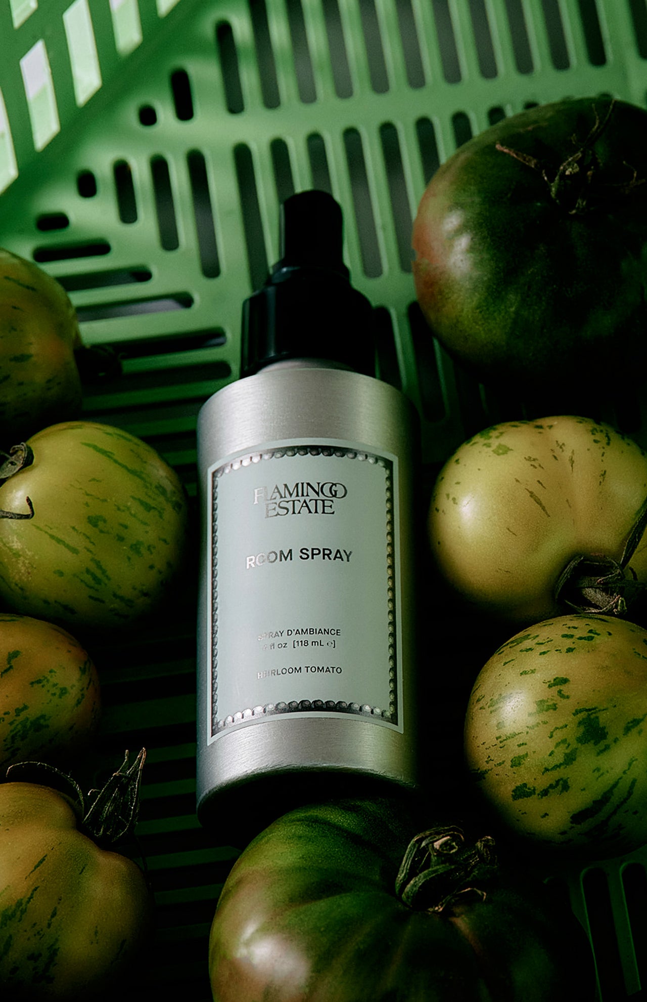 A silver bottle labeled “FLAMINGO ESTATE Roma Heirloom Tomato Room Spray” rests among green and yellow heirloom tomatoes in a green basket, evoking the essence of this room spray that captures the fresh, summer garden scent for your home fragrance collection.