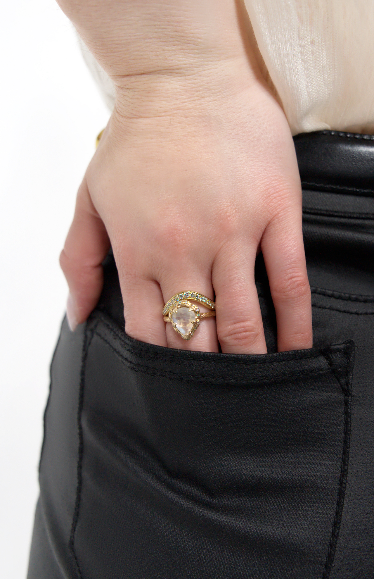 A hand with a LOGAN HOLLOWELL Enigma Channel Band Ring in yellow gold rests on the back pocket of black pants, paired with a cream-colored top—showcasing refined designer jewelry style.