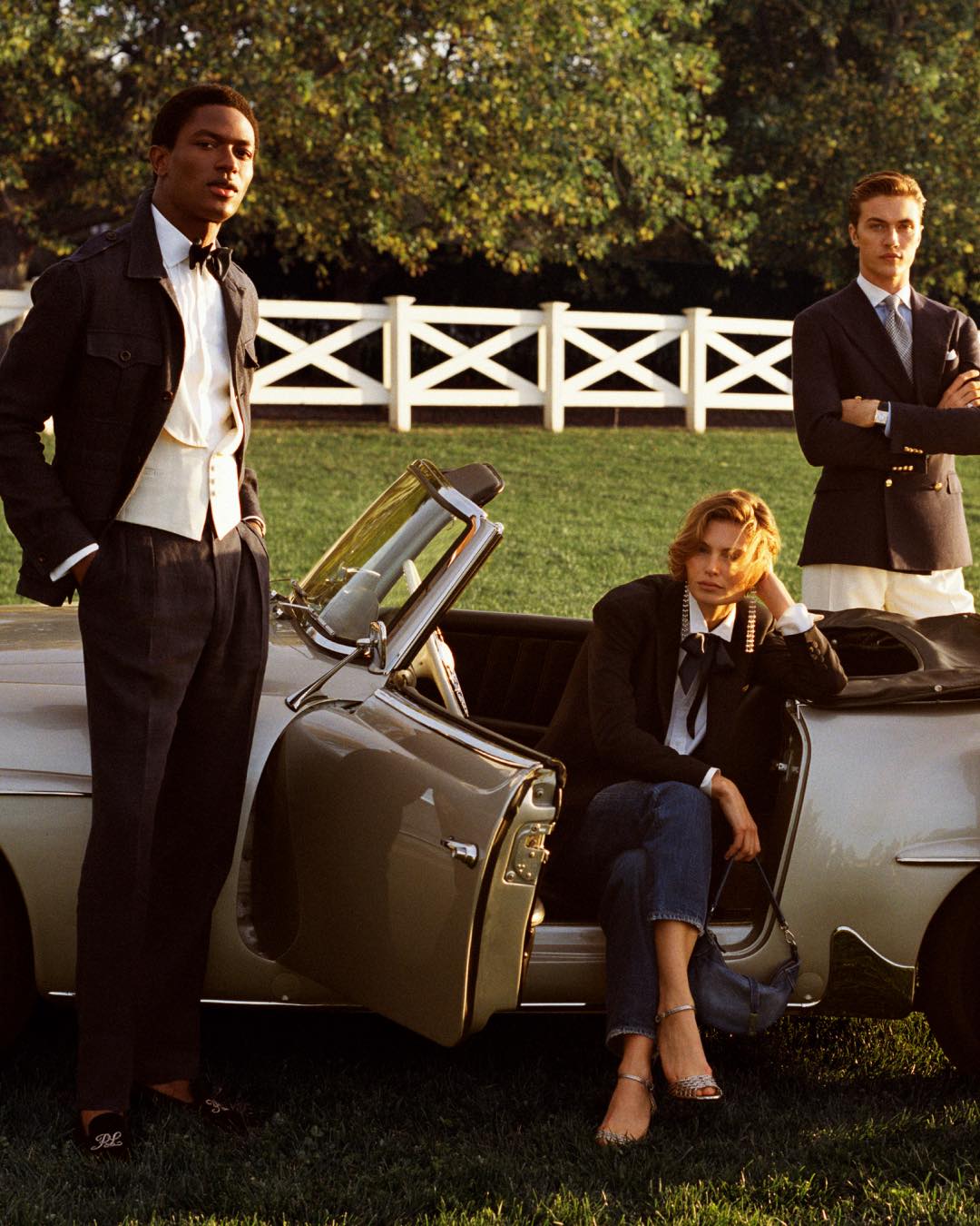 Three people in formal attire standing around a vintage car in a grassy area with a white fence and trees in the background.