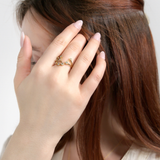 A woman with long brown hair covers part of her face, displaying the delicate LOGAN HOLLOWELL Aquarius Constellation Ring on her hand. She has light pink nails, wears a cream top and gold necklace, set against a plain light background.