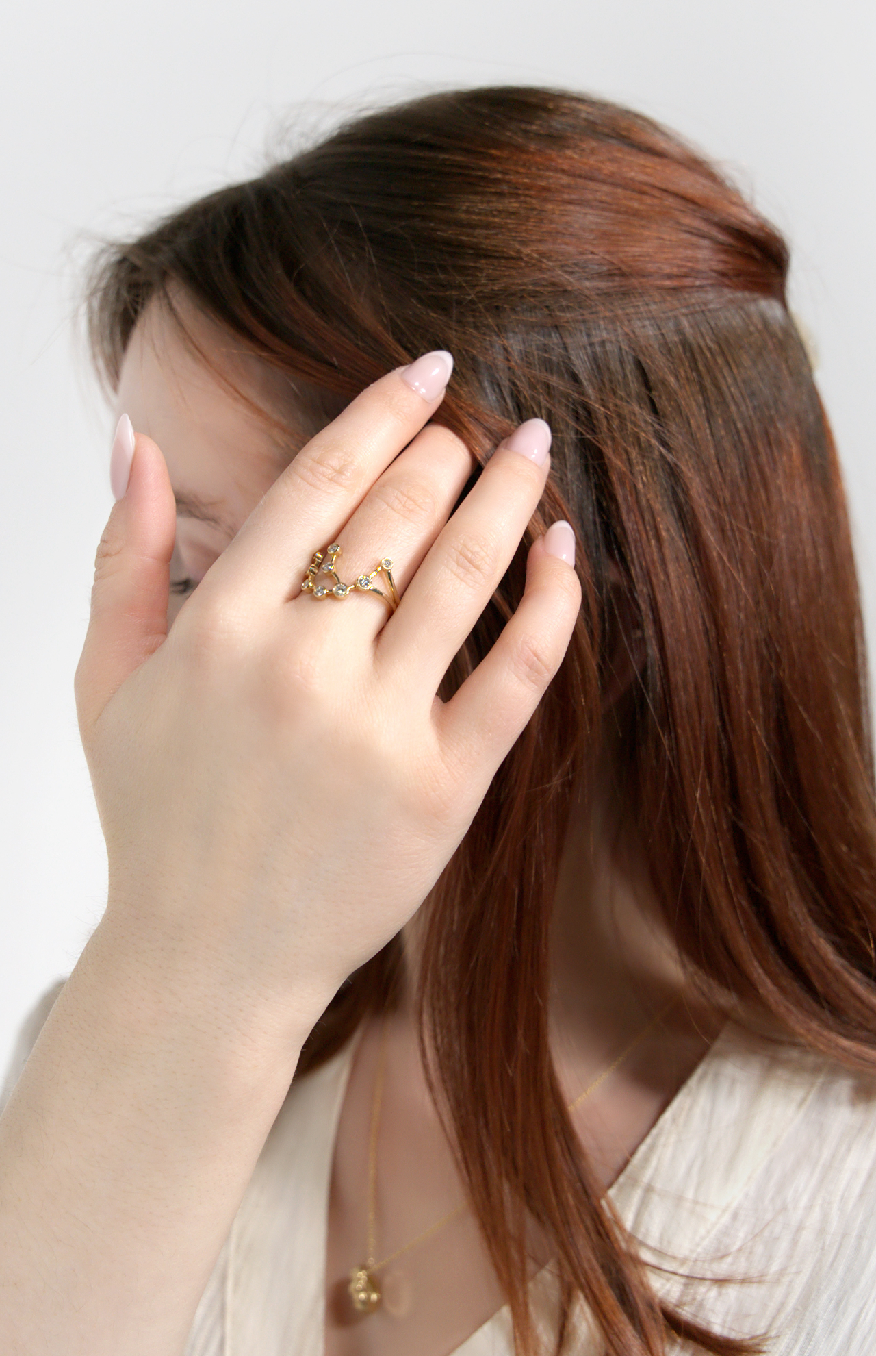 A woman with long brown hair covers part of her face, displaying the delicate LOGAN HOLLOWELL Aquarius Constellation Ring on her hand. She has light pink nails, wears a cream top and gold necklace, set against a plain light background.