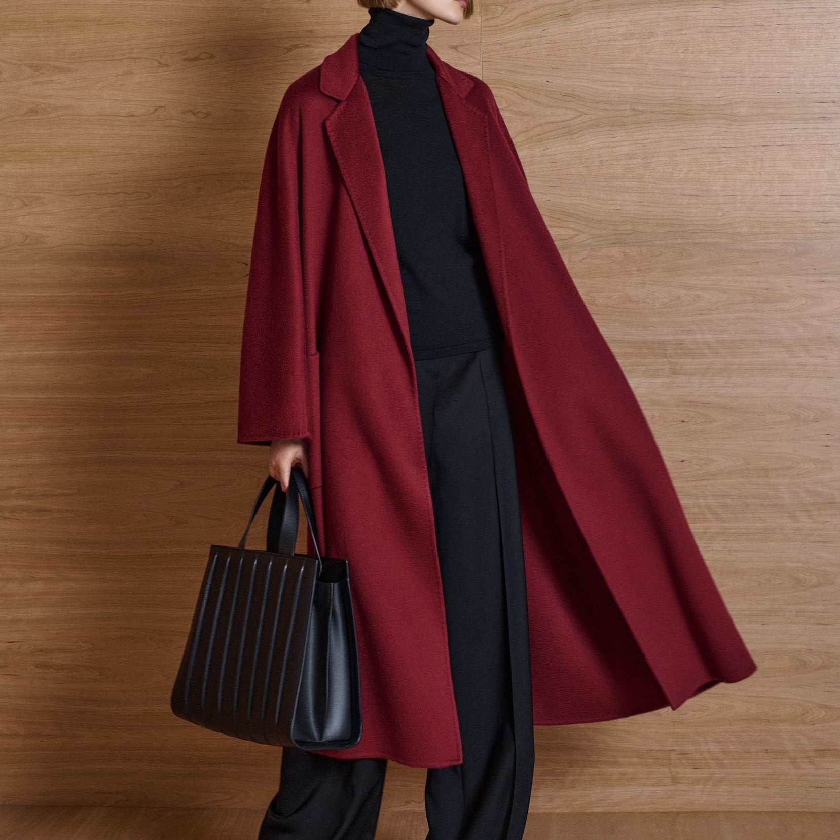 Person wearing a long red coat and black pants, holding a black handbag against a wooden background