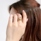 A woman with brown hair showcases a LOGAN HOLLOWELL Enigma Channel Band Ring, featuring yellow gold with a large pear-shaped stone and matching diamond band. Her light pink manicured nails complete the elegant look.