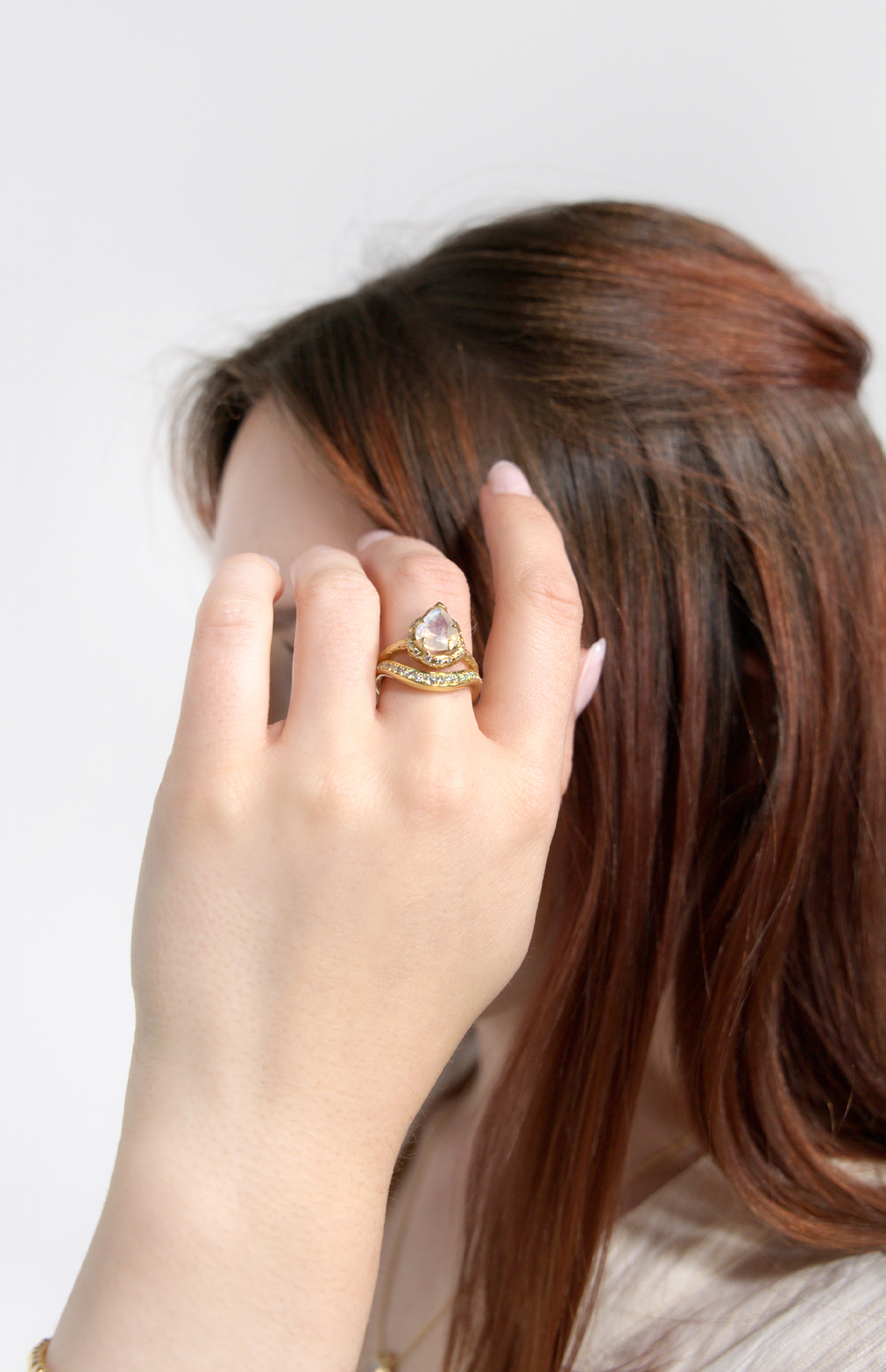 A woman with brown hair showcases a LOGAN HOLLOWELL Enigma Channel Band Ring, featuring yellow gold with a large pear-shaped stone and matching diamond band. Her light pink manicured nails complete the elegant look.