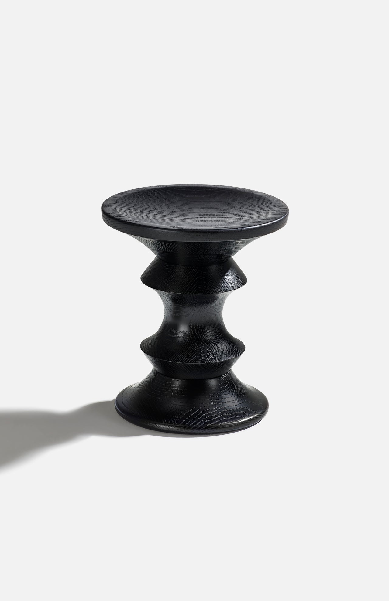 The HERMAN MILLER Eames Turned Stool, Shape A, features a black finish with a round top and base, three wavy concave midsections in solid walnut style, and casts a leftward shadow on a plain light background.