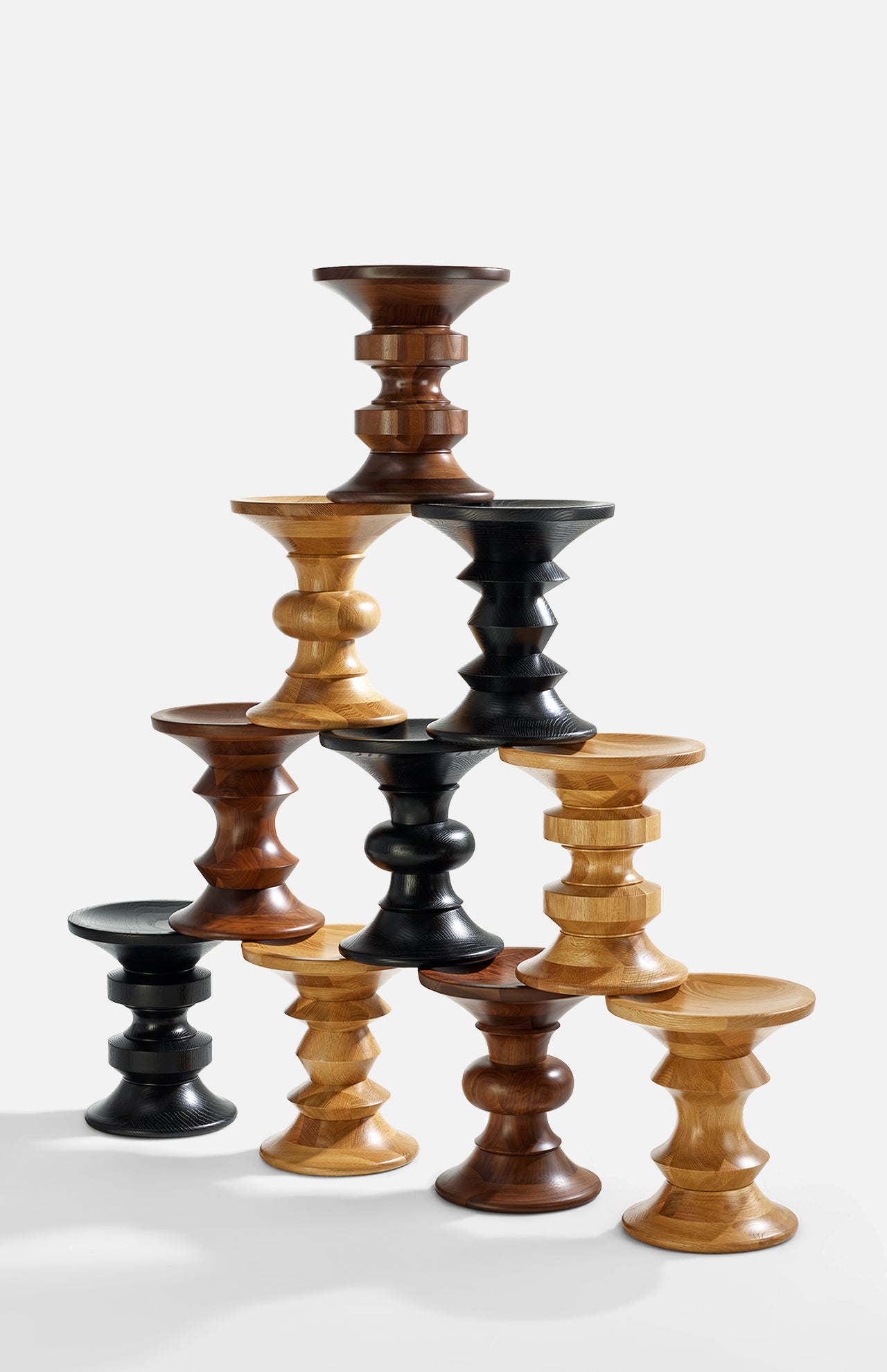 Seven Eames Turned Stools, Shape D by Herman Miller are stacked in a pyramid, showcasing their solid walnut and lighter finishes against a plain white background.