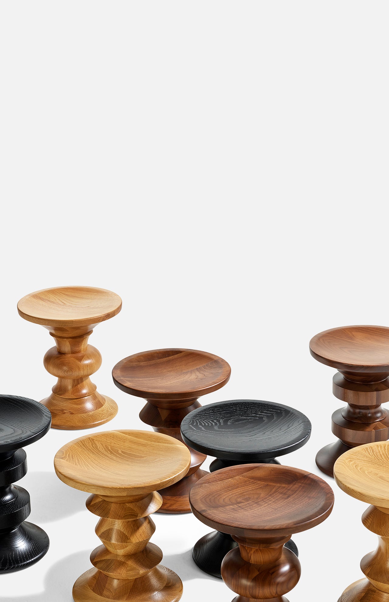 A cluster of HERMAN MILLER Eames Turned Stool, Shape D in solid walnut and other wood finishes—light, medium, dark brown, and black—stands sculpturally against a plain white background.