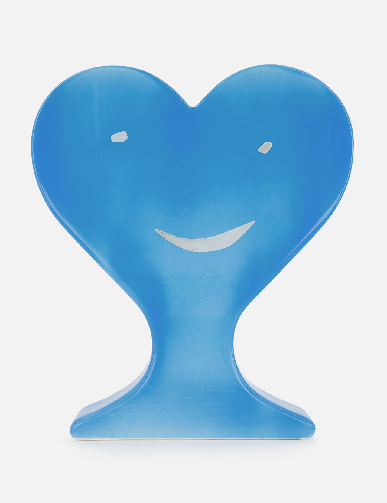 A blue, heart-shaped sculpture from TOT COR’s Corazones by Jordi Torres features a simple smiling face—two dot eyes and a curved mouth—standing upright on a flat base against a plain white background.