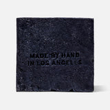 The FLAMINGO ESTATE Lavender Activated Charcoal Soap Brick features a rough texture, is stamped MADE BY HAND IN LOS ANGELES on the front, and appears against a plain white background for an aromatic cleansing experience.