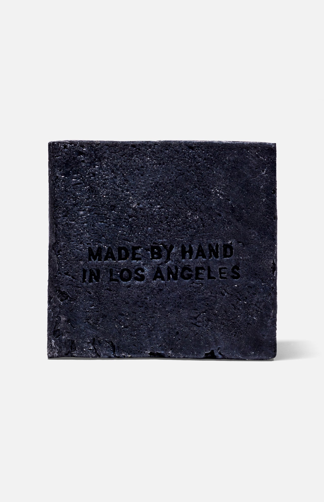 The FLAMINGO ESTATE Lavender Activated Charcoal Soap Brick features a rough texture, is stamped MADE BY HAND IN LOS ANGELES on the front, and appears against a plain white background for an aromatic cleansing experience.