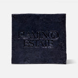 A black, textured square plaque with "FLAMINGO ESTATE" embossed at the center, reminiscent of the Lavender Activated Charcoal Soap Brick by FLAMINGO ESTATE, displayed on a plain white background.