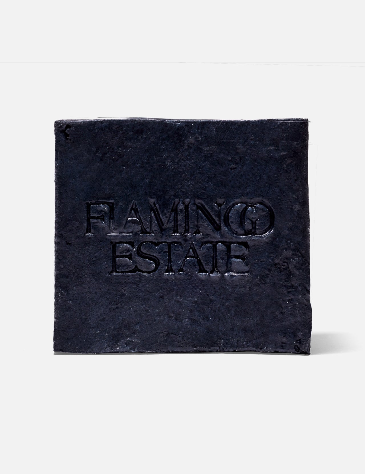 A black, textured square plaque with "FLAMINGO ESTATE" embossed at the center, reminiscent of the Lavender Activated Charcoal Soap Brick by FLAMINGO ESTATE, displayed on a plain white background.