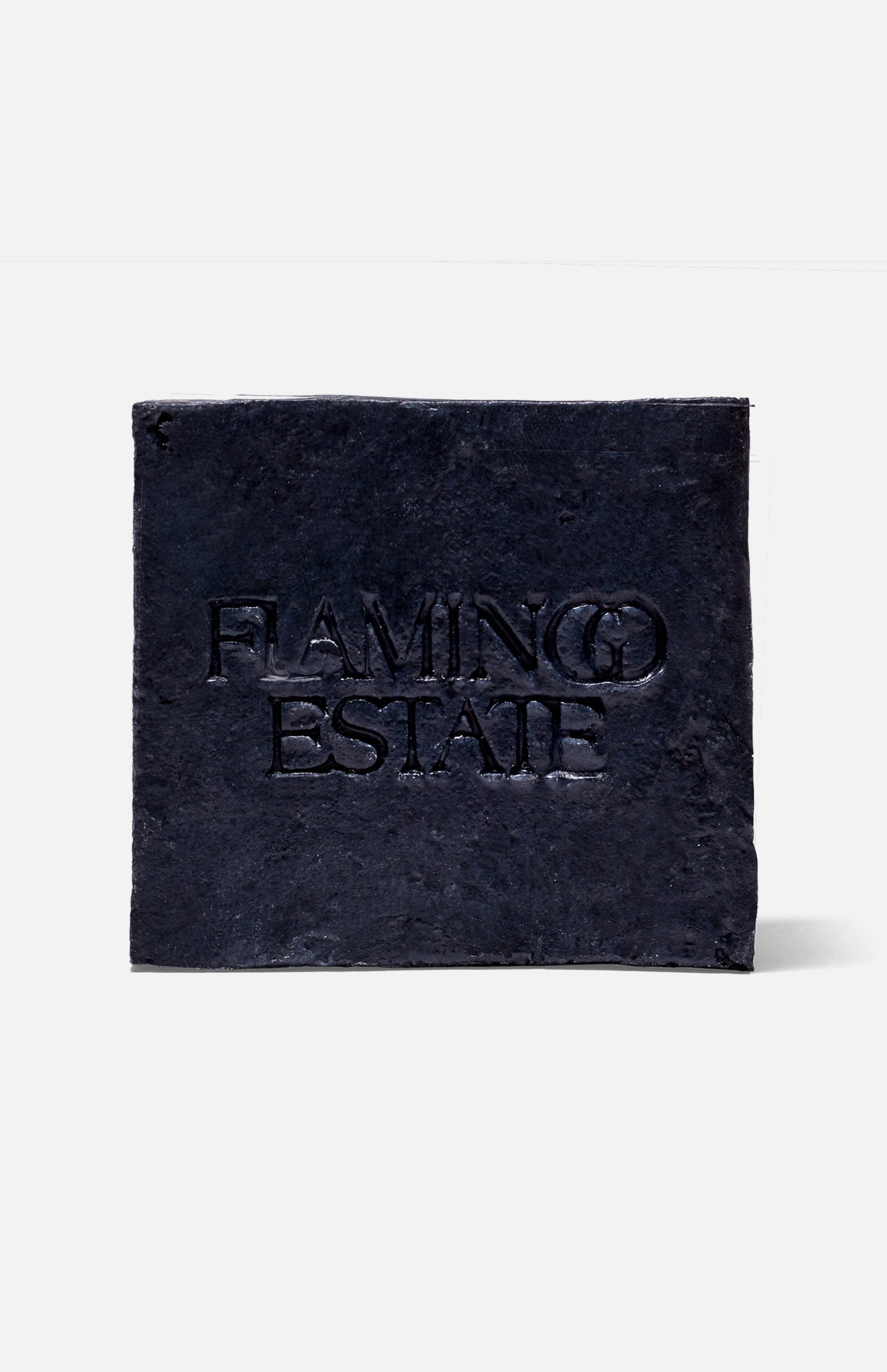 A black, textured square plaque with "FLAMINGO ESTATE" embossed at the center, reminiscent of the Lavender Activated Charcoal Soap Brick by FLAMINGO ESTATE, displayed on a plain white background.