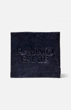 A black, textured square plaque with "FLAMINGO ESTATE" embossed at the center, reminiscent of the Lavender Activated Charcoal Soap Brick by FLAMINGO ESTATE, displayed on a plain white background.